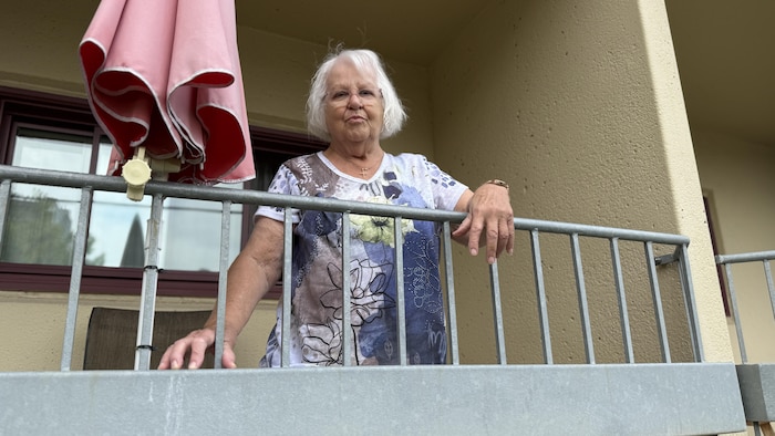 Une femme sur un balcon, la main gauche appuyée contre les barreaux, regarde la caméra.