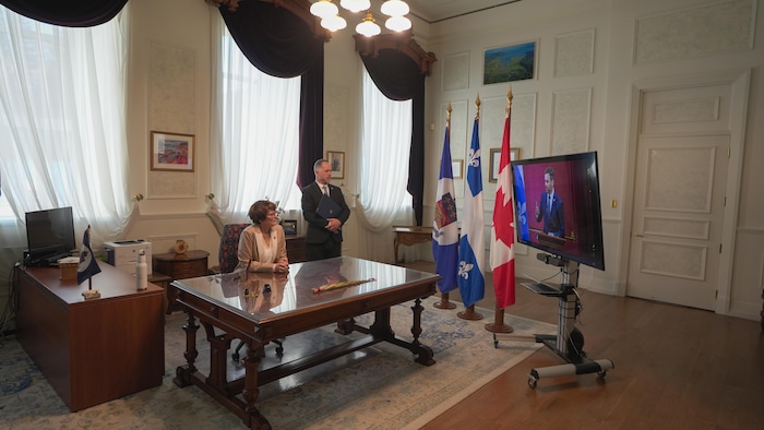 Manon Jeannotte, assise à son bureau, regarde la télévision.