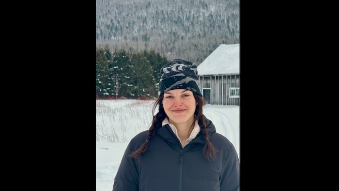 Elle est devant un flan de montagne et devant des sapins et un bâtiment agricole.