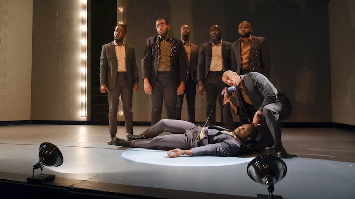 Seven actors on stage, one lying on the floor and another kneeling next to him, speaking into a microphone. 