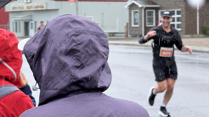 Un homme portant un dossard, courant sous la pluie. À l'avant plan, des témoins soutiennent les coureurs.