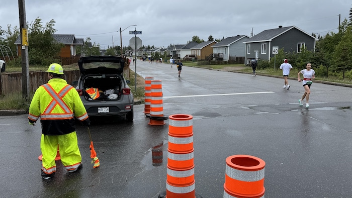 Un homme en habits de construction portant un drapeau derrière des cônes alors que des gens courent dans la rue.