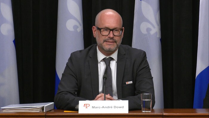 Marc-André Dowd en conférence de presse, devant des drapeaux du Québec.