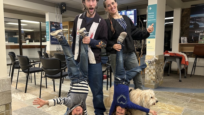 Deux adultes sont côte à côte et tiennent chacun la jambe d'un enfant qui a la tête en bas. Ils sourient tous et regardent la caméra. 