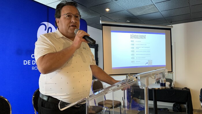 Marc Bibeau debout dans une salle, microphone en main, parle à son auditoire. 