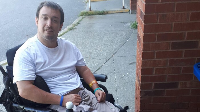 Marc Carroll est assis sur un fauteuil roulant et porte un chandail, un pantalon et des bracelets.