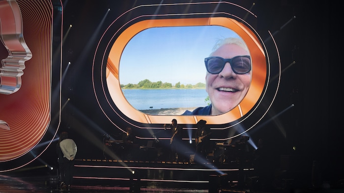 L'animateur apparaît dans une vidéo tournée à l'extérieur, projetée sur la scène. 