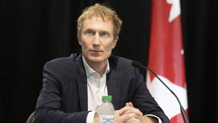 مارك ميلر جالساً في مؤتمر صحفي وخلفه علم كندا.