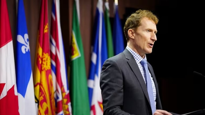 Marc Miller en conférence de presse à Ottawa, devant les drapeaux du Canada et des provinces et territoires.