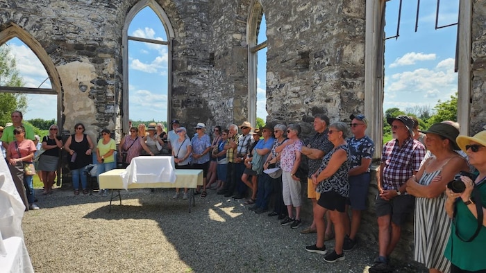 Des gens se tiennent debout à l'intérieur le long des murs intérieurs de l'église qui n'a plus de toit ni de fenêtres.