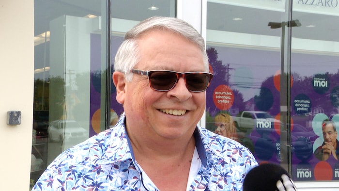 Un homme aux cheveux blancs courts portant des lunettes fumées et une chemise estivale est dehors devant une pharmacie.