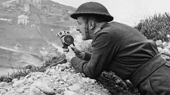 Juché sur une butte sur le front italien, le correspondant de guerre Marcel Ouimet, portant un casque militaire, enregistre un
reportage un micro à la main.