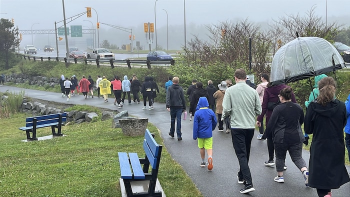 Plusieurs personnes marchant sur un sentier bordant une autoroute sous la pluie.