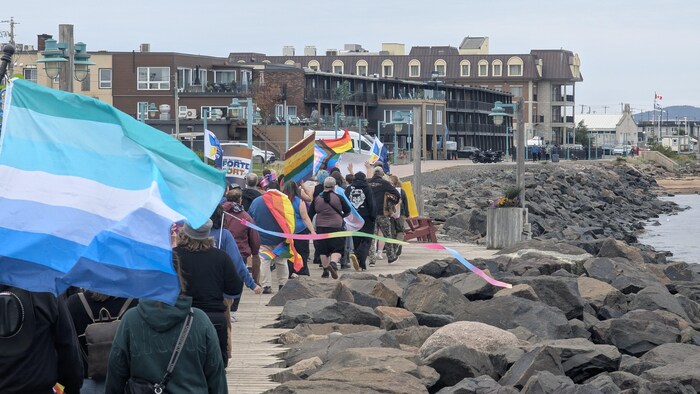 Un groupe de personnes de dos marche sur une promenade en tenant de grands drapeaux arc-en-ciel et d'autres drapeaux colorés.
