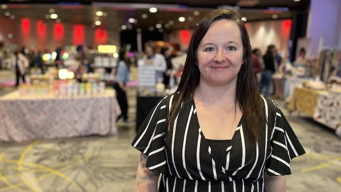 Une femme pose devant une grande salle où sont rassemblés de nombreux artisans.