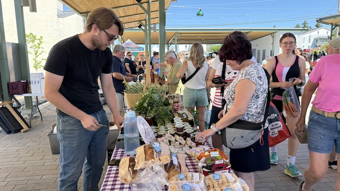 William Pack, derrière ses produits disposés sur un comptoir dans un kiosque, avec une cliente qui regarde les produits.