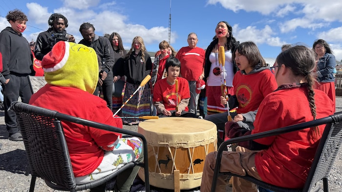 Des enfants, tous vêtus de rouge, sont en cercle autour d'un tambour traditionnel. Ils ont tous une baguette pour frapper sur le tambour. Un des enfants chante, accompagné d'une femme derrière avec une main rouge en peinture sur son visage.