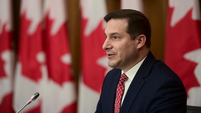 Le ministre de l'Immigration, vêtu d'un veston et d'une cravate, parle dans un micro devant des drapeaux du Canada.