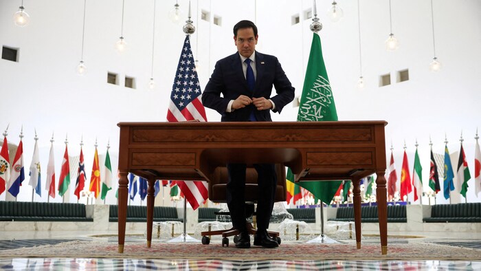 Un homme attache son veston devant une table entourée de drapeaux.