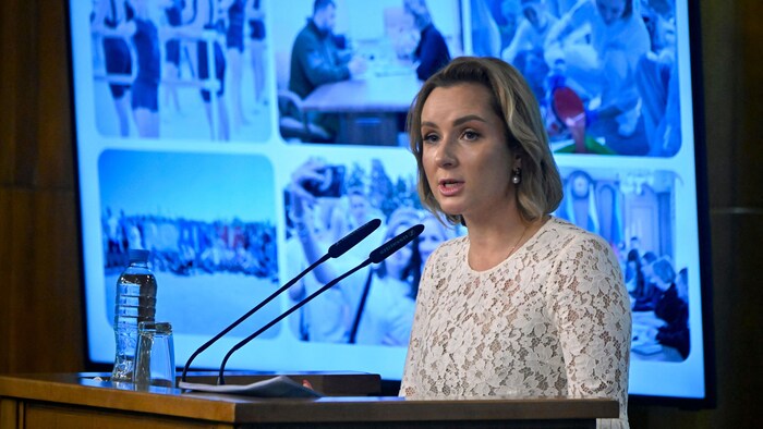 Maria Lavova-Belova mavi ekranın önünde mikrofona konuşuyor. 