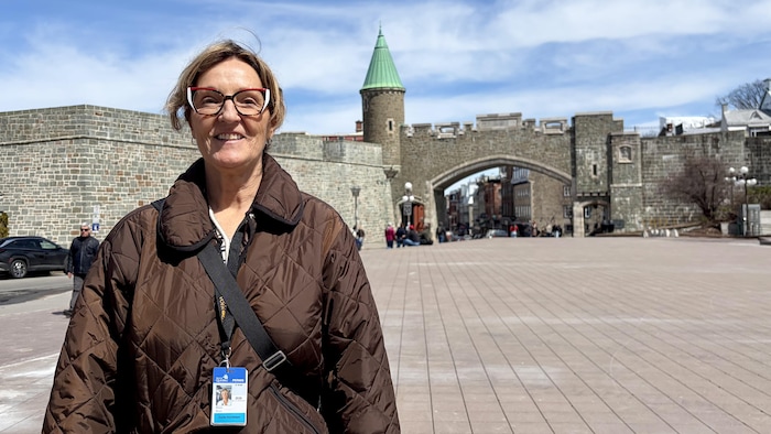 Marie Blais sourit à la caméra, sur la place d'Youville. En arrière-plan, les fortifications de Québec et la porte Saint-Jean. 