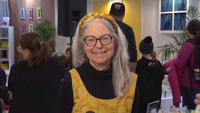 Une femme souriante habillée en jaune et noir, devant un kiosque fréquenté par plusieurs passants.