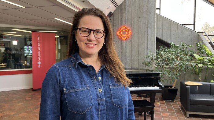 Une femme souriante à lunettes pose dans un studio de Radio-Canada. Derrière elle se trouvent un piano à queue noir, le logo néon rouge de la SRC, une bannière « Saguenay–Lac-Saint-Jean » et des plantes vertes d'intérieur.