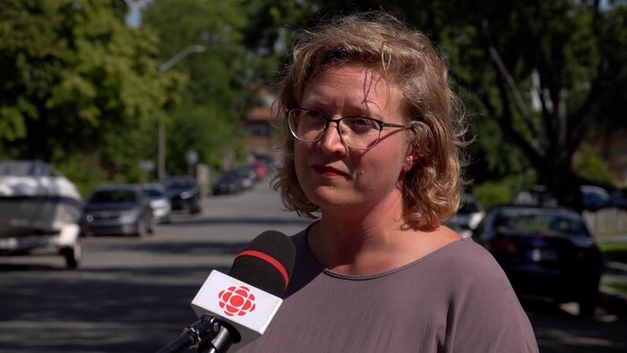 Elle donne une entrevue au micro de Radio-Canada dans une rue avec des arbres en arrière-plan.