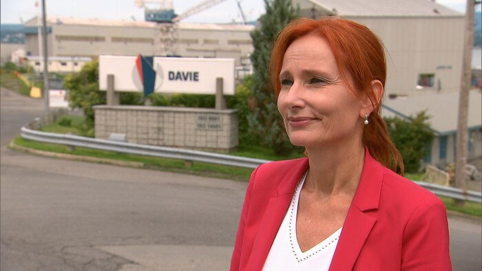 Marie-Josée Morency en entrevue, à l'extérieur, devant l'entrée du Chantier Davie. Elle porte un veston rouge. Elle a un regard fier.