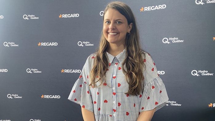 Une femme souriante aux longs cheveux châtains pose devant un mur de commanditaires arborant les logos « REGARD » et « Hydro-Québec ». Elle porte une robe boutonnée à rayures verticales bleues et blanches, parsemée de petits cœurs rouges.