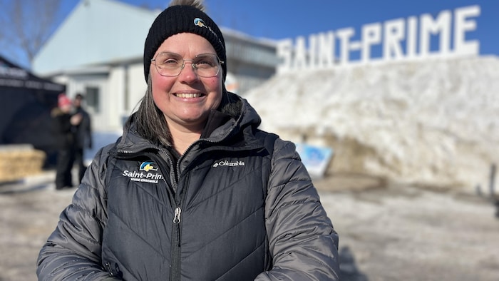 Une femme souriante portant des vêtements d'hiver et une tuque noire pose devant un grand panneau indiquant « SAINT-PRIME ». L'arrière-plan montre une butte de neige, un bâtiment gris et un ciel bleu clair sous un soleil éclatant.