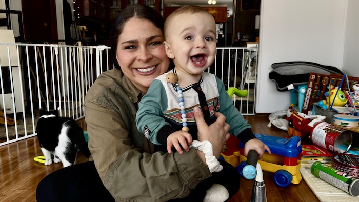 Une femme et son bébé sont assis par terre près de jouets et d’un chat et sourient à la caméra.