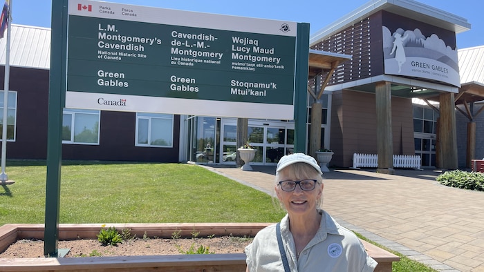 Marie Walsh est debout devant le panneau d'entrée du site patrimonial de Green Gables. Elle sourit à la caméra.