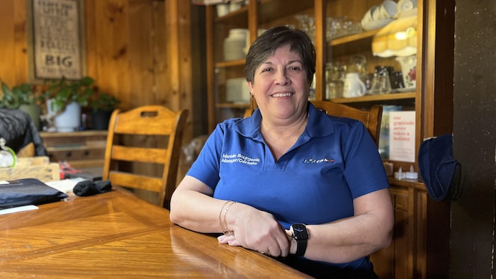 Une femme sourit, assise à une table en bois, devant un vaisselier.
