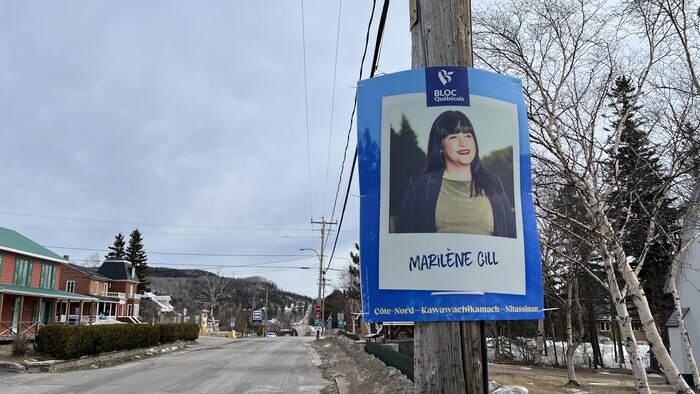 Une pancarte électorale est accrochée à un poteau en bordure de route. Une rue et des maisons sont visibles derrière. 