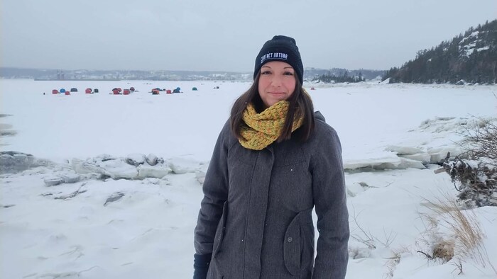 Une femme portant un manteau gris, un foulard jaune et une tuque noire sourit devant un plan d'eau gelé. À l'arrière-plan, plusieurs tentes de pêche colorées sont dispersées sur la glace sous un ciel couvert et hivernal.