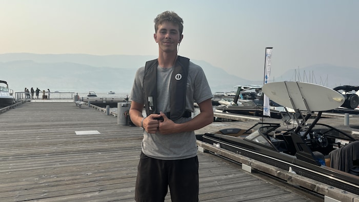 Un jeune homme aux cheveux blonds bouclés, portant un t-shirt gris et un gilet sombre, se tient debout sur un quai de bois. Autour de lui, des embarcations de plaisance amarrées.