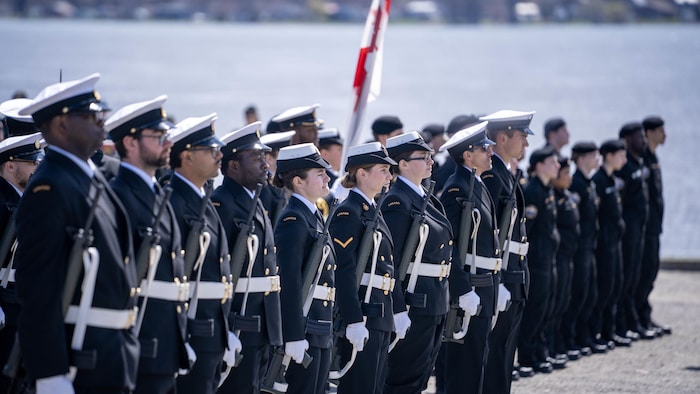 Des membres de la Marine royale canadienne en uniforme et installés en rang avec armes et drapeaux à la main. 
