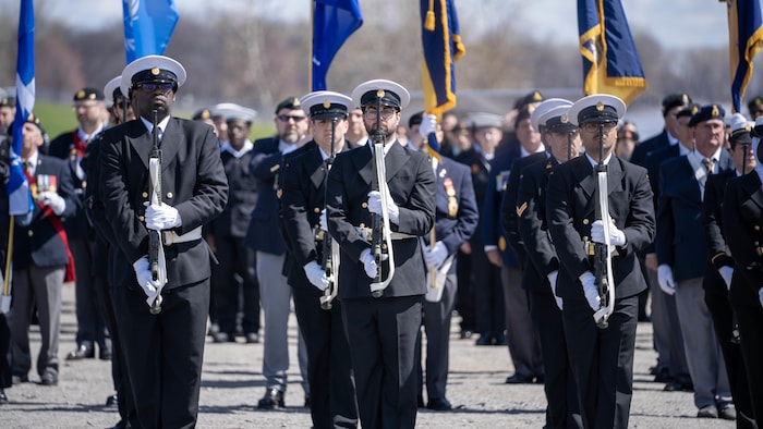 Des membres de la Marine royale canadienne en uniforme sont rassemblés sur le bord du fleuve à Trois-Rivières avec des drapeaux à la main. 