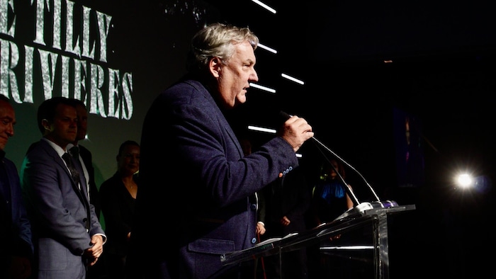 Mario De Tilly parle dans un micro sur une scène.                              