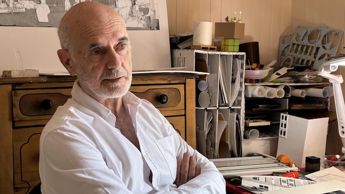 Mario Dufour est dans son atelier, assis à un bureau où se trouvent des quantités de papiers, de documents et d'outils.