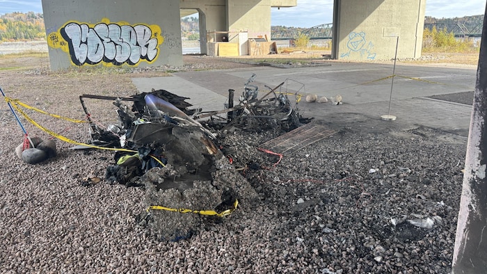 Des débris calcinés gisent au sol sur du gravier, entourés d'un ruban de sécurité jaune sous les structures en béton d'un pont.
