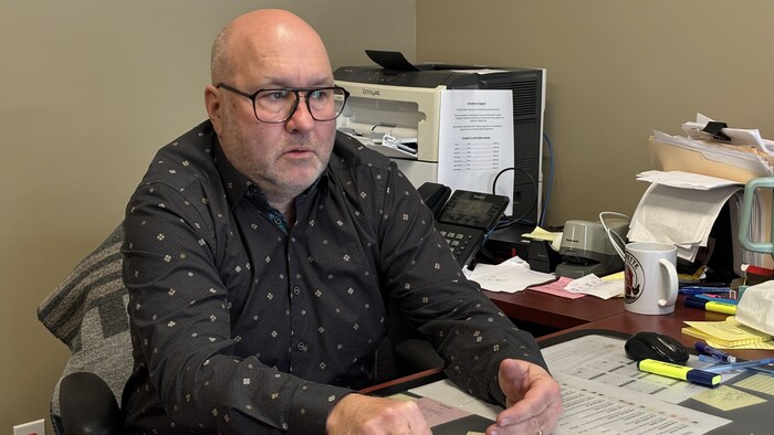 Un homme portant des lunettes est assis derrière un bureau, près d’un ordinateur.
