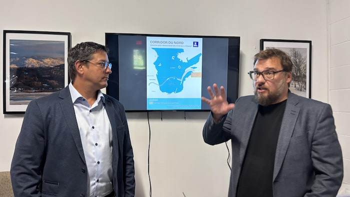 Deux hommes en complet, dont l'un d'eux est en train de parler, la main levée, devant une carte affichée sur un écran.