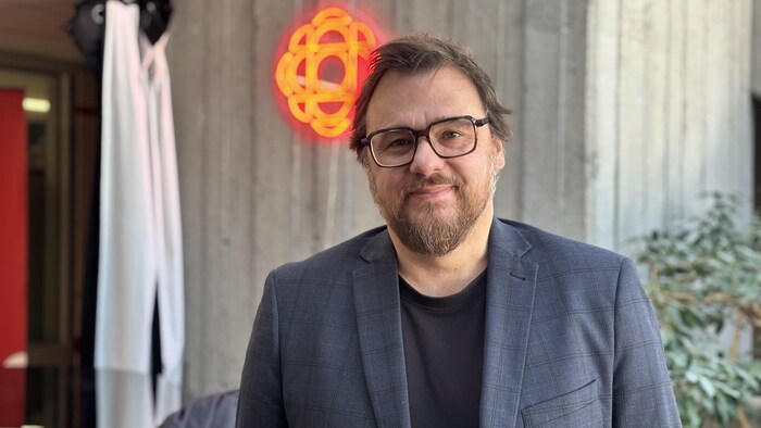 Un homme d'âge moyen à lunettes et barbe courte sourit devant un mur de béton. Il porte un complet à carreaux sur un chandail noir. En arrière-plan, un logo lumineux rouge de Radio-Canada est visible.