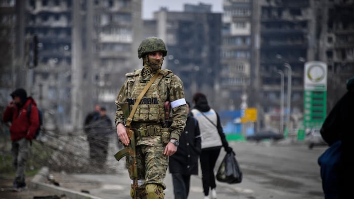 Un soldat russe au milieu d'une rue. En arrière-plan, des gens marchent dans une rue. Des immeubles de logements carbonisés sont visibles. 