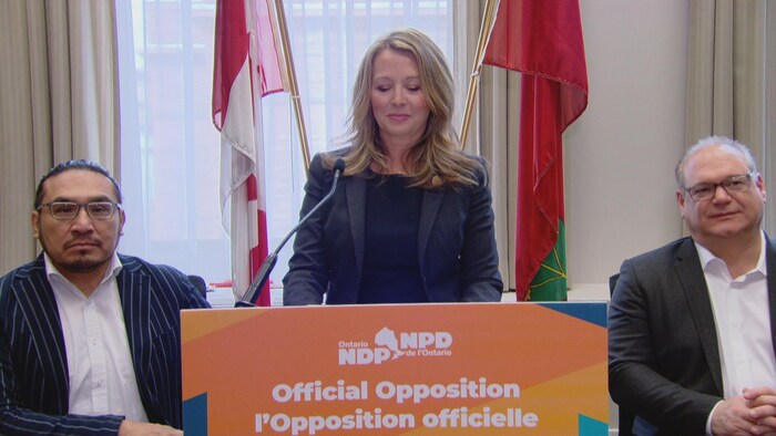 Marit Stiles, debout derrière un podium orange, entourée de deux membres de son caucus.