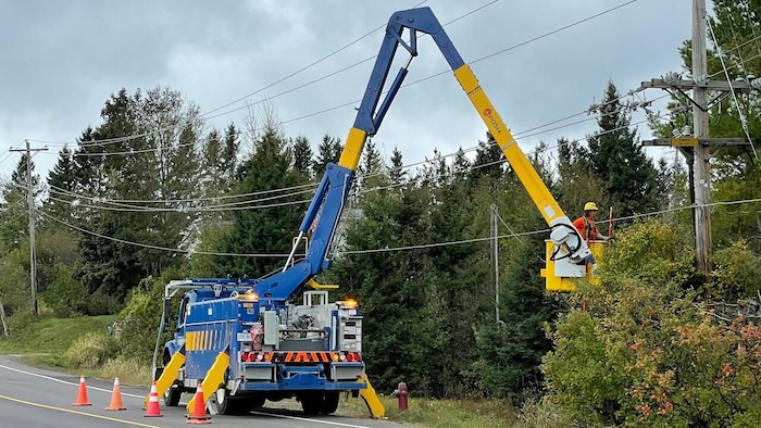 Un camion bleu immobilisé sur une route. Un homme dans une nacelle travaille sur des lignes de transmission électrique.