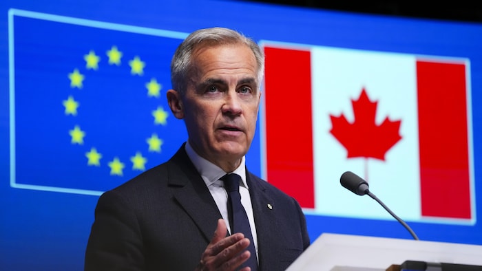 Mark Carney parle dans un micro lors d'une conférence de presse, devant un écran affichant les drapeaux du Canada et de l'Union européenne.