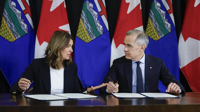 Mark Carney et Danielle Smith sont assis et signent des documents.
